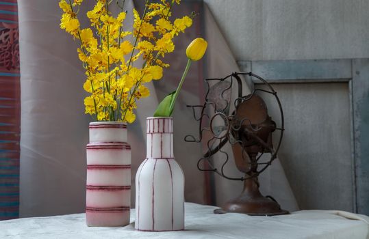 Bouquet of yellow flowers in Two handmade ceramic vase and Vintage fan on white textured table cloth with old cement wall. Home decor, Selective focus.