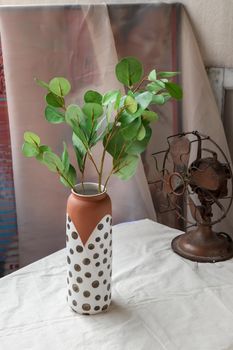 Green leaf in Handmade ceramic vase and Vintage fan on white textured table cloth with old cement wall. Home decor, Selective focus.