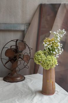 Bouquet of green and white flowers in Handmade ceramic vase and Vintage fan on white textured table cloth with old cement wall. Home decor, Selective focus.