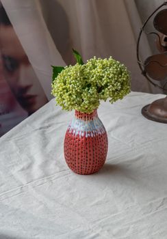 Bangkok, Thailand - Feb 06, 2021 : Bouquet of green flowers in Red handmade ceramic vase and old vintage fan on white textured table cloth in front of Classic chinese poster movie frame with old cement wall. Home decor, Selective focus.