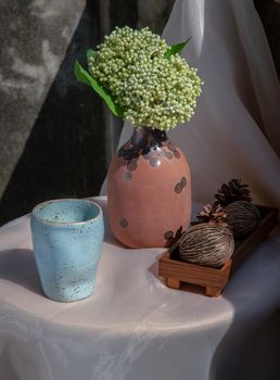 Blue ceramic mug handmade with bunch of flowers in vase and Dried cerbera oddloam's on white table in vintage room with old ruins cement wall. Selective focus.