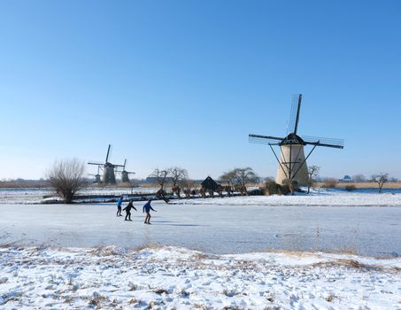 people skate on the ice of canal near kinderdijk with a lot of windmills in holland on sunny winter day