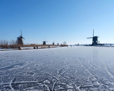 people skate on the ice of canal near kinderdijk with a lot of windmills in holland on sunny winter day