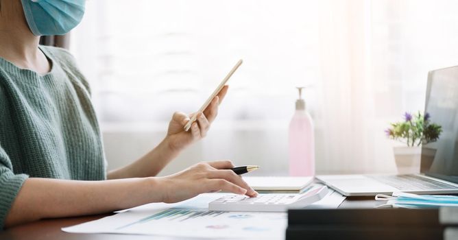 accounting, taxes and finances concept - woman with smartphone and calculator wearing face protective medical mask for protection from virus disease at home