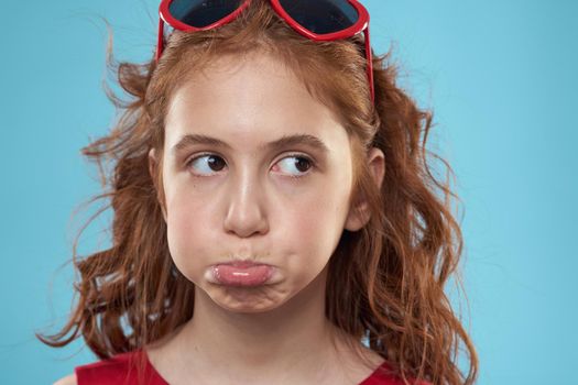 Little girl in sunglasses in the form of hearts red dress curly hair blue background. High quality photo