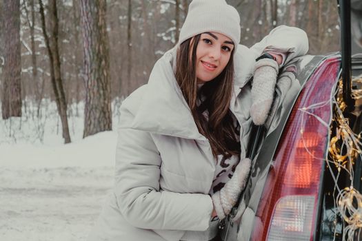 Attractive beautiful young woman in sweater staying next to the car in forest. Winter vacation. Holidays. Smilling Christmas lights.