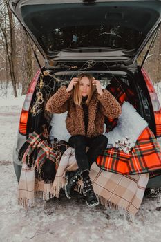 Blonde woman wrapped in blanket in trunk car. Travel in winter. Car decorated with festive Christmas lights. Outdoor picnic. Unity with nature