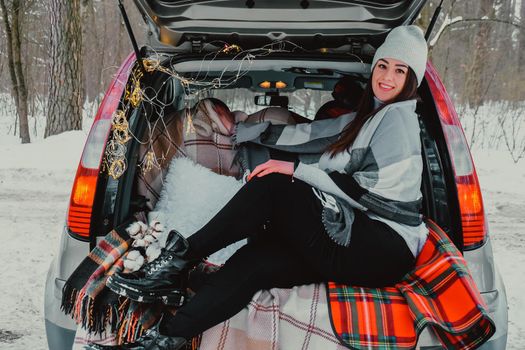 Brunette woman wrapped in blanket in trunk car. Travel in winter. Car decorated with festive Christmas lights. Outdoor picnic. Unity with nature