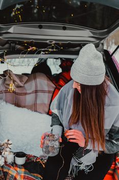 Brunette woman wrapped in blanket in trunk car. Travel in winter. Car decorated with festive Christmas lights. Outdoor picnic. Unity with nature