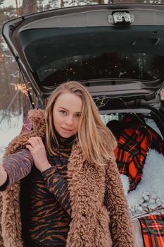 Attractive beautiful young woman in sweater staying next to the car in forest. Winter vacation. Holidays. Smilling Christmas lights.