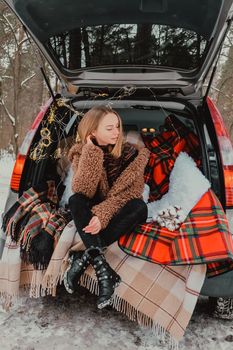 Blonde woman wrapped in blanket in trunk car. Travel in winter. Car decorated with festive Christmas lights. Outdoor picnic. Unity with nature