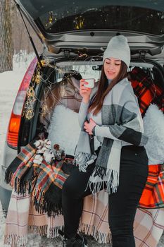 Brunette woman wrapped in blanket in trunk car drinking coffee tea from thermos. Travel in winter. Car decorated with festive Christmas lights. Outdoor picnic. Unity with nature