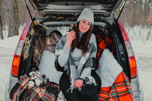 Brunette woman wrapped in blanket in trunk car drinking coffee tea from thermos. Travel in winter. Car decorated with festive Christmas lights. Outdoor picnic. Unity with nature