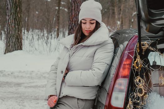 Attractive beautiful young woman in sweater staying next to the car in forest. Winter vacation. Holidays. Smilling Christmas lights.