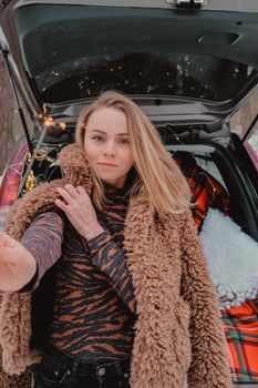 Attractive beautiful young woman in sweater staying next to the car in forest. Winter vacation. Holidays. Smilling Christmas lights.