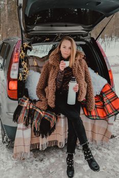 Blonde woman wrapped in blanket in trunk car drinking coffee tea from thermos. Travel in winter. Car decorated with festive Christmas lights. Outdoor picnic. Unity with nature