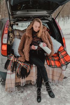 Blonde woman wrapped in blanket in trunk car drinking coffee tea from thermos. Travel in winter. Car decorated with festive Christmas lights. Outdoor picnic. Unity with nature