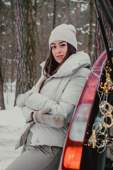 Attractive beautiful young woman in sweater staying next to the car in forest. Winter vacation. Holidays. Smilling Christmas lights.