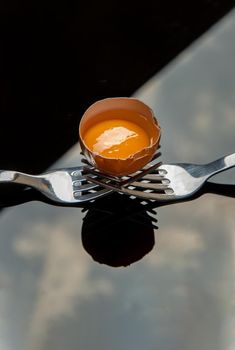 Broken fresh chicken egg (Hen egg) balanced on a composition of two intertwined forks over black table.  Copy space, Selective focus.