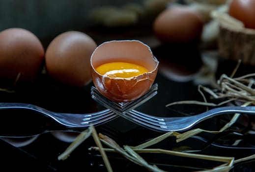 Close-up of Broken fresh chicken egg (Hen egg) balanced on a composition of two intertwined forks with fresh chicken eggs on wood background. Selective focus.