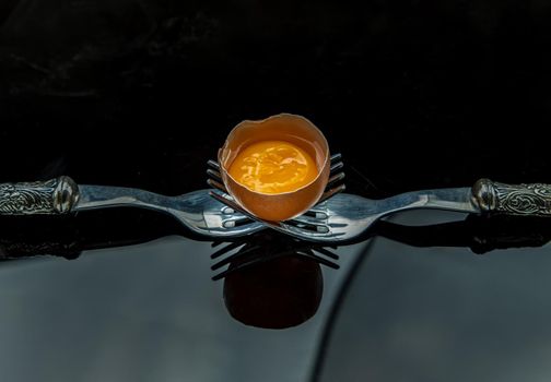 Broken fresh chicken egg (Hen egg) balanced on a composition of two intertwined forks over black table. Copy space, Selective focus.