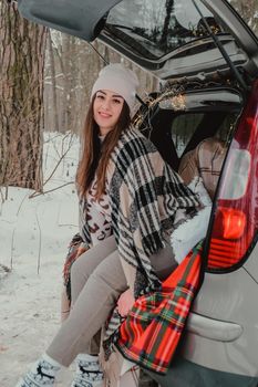 Brunette woman wrapped in blanket in trunk car drinking coffee tea from thermos. Travel in winter. Car decorated with festive Christmas lights. Outdoor picnic. Unity with nature