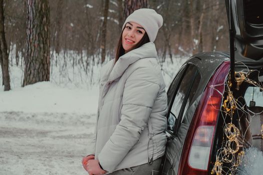 Attractive beautiful young woman in sweater staying next to the car in forest. Winter vacation. Holidays. Smilling Christmas lights.