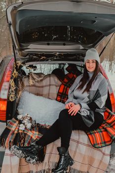 Brunette woman wrapped in blanket in trunk car. Travel in winter. Car decorated with festive Christmas lights. Outdoor picnic. Unity with nature