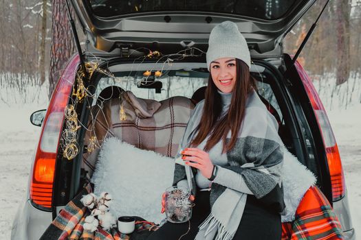 Brunette woman wrapped in blanket in trunk car. Travel in winter. Car decorated with festive Christmas lights. Outdoor picnic. Unity with nature