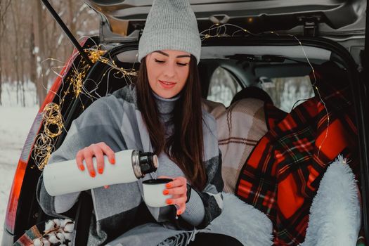 Brunette woman wrapped in blanket in trunk car drinking coffee tea from thermos. Travel in winter. Car decorated with festive Christmas lights. Outdoor picnic. Unity with nature