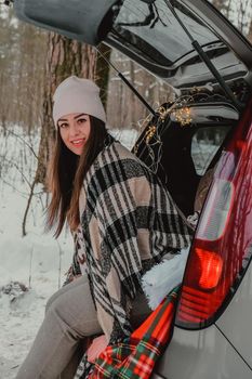 Brunette woman wrapped in blanket in trunk car drinking coffee tea from thermos. Travel in winter. Car decorated with festive Christmas lights. Outdoor picnic. Unity with nature