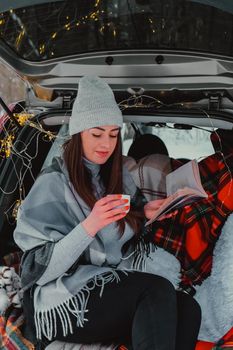 Brunette woman wrapped in blanket in trunk car drinking coffee tea from thermos. Travel in winter. Car decorated with festive Christmas lights. Outdoor picnic. Unity with nature