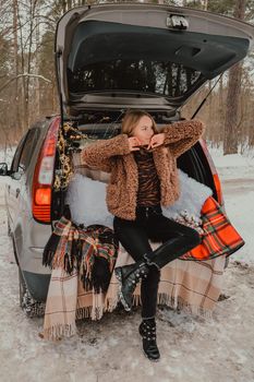 Blonde woman wrapped in blanket in trunk car. Travel in winter. Car decorated with festive Christmas lights. Outdoor picnic. Unity with nature