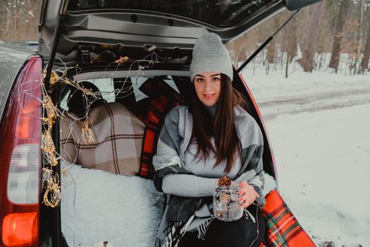 Brunette woman wrapped in blanket in trunk car. Travel in winter. Car decorated with festive Christmas lights. Outdoor picnic. Unity with nature