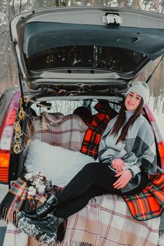 Brunette woman wrapped in blanket in trunk car. Travel in winter. Car decorated with festive Christmas lights. Outdoor picnic. Unity with nature