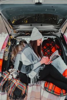 Brunette woman wrapped in blanket in trunk car drinking coffee tea from thermos. Travel in winter. Car decorated with festive Christmas lights. Outdoor picnic. Unity with nature