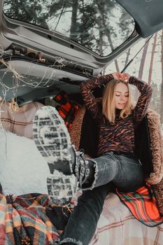 Blonde woman wrapped in blanket in trunk car. Travel in winter. Car decorated with festive Christmas lights. Outdoor picnic. Unity with nature