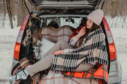 Brunette woman wrapped in blanket in trunk car drinking coffee tea from thermos. Travel in winter. Car decorated with festive Christmas lights. Outdoor picnic. Unity with nature