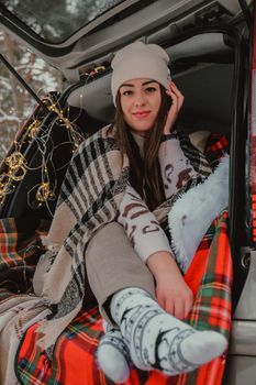 Brunette woman wrapped in blanket in trunk car drinking coffee tea from thermos. Travel in winter. Car decorated with festive Christmas lights. Outdoor picnic. Unity with nature