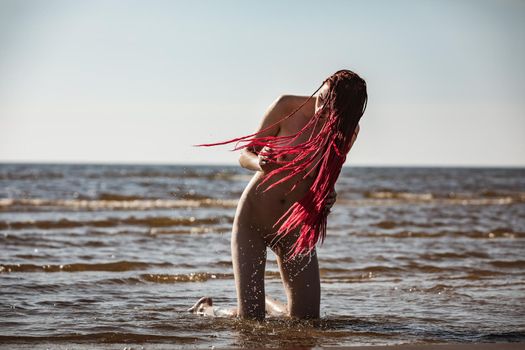 Beautiful girl outdoors enjoying nature. Young naked woman with scarlet dreadlocks enjoys the sea on the coast