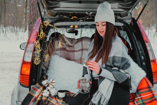 Brunette woman wrapped in blanket in trunk car. Travel in winter. Car decorated with festive Christmas lights. Outdoor picnic. Unity with nature