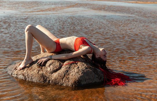 Beautiful girl outdoors enjoying nature. Seminude girl with scarlet dreadlocks in a red bathing suit sunbathes on the beach