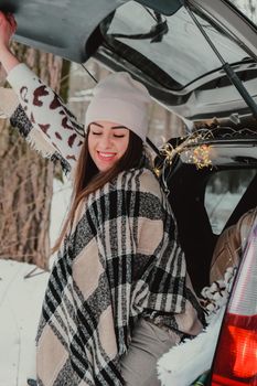 Brunette woman wrapped in blanket in trunk car drinking coffee tea from thermos. Travel in winter. Car decorated with festive Christmas lights. Outdoor picnic. Unity with nature