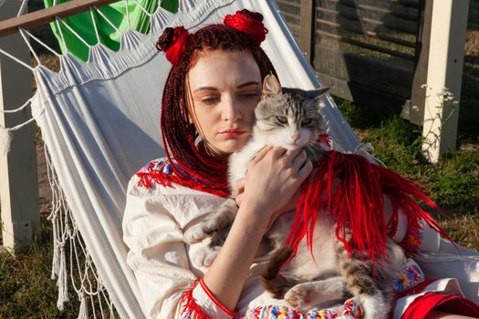 Young woman with scarlet dreadlocks in national dress lying in a hammock and playing with a cat. Outdoors portrait