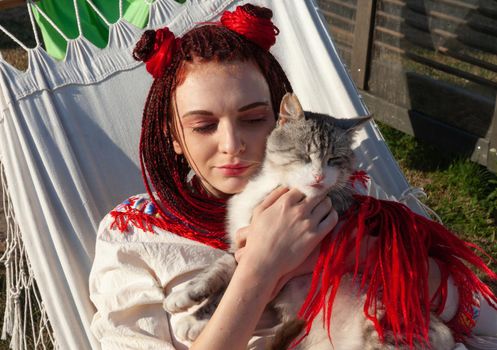 Young woman with scarlet dreadlocks in national dress lying in a hammock and playing with a cat. Outdoors portrait