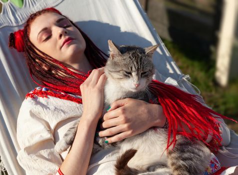 Young woman with scarlet dreadlocks in national dress lying in a hammock and playing with a cat. Outdoors portrait. Focus on the cat