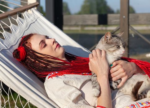 Young woman with scarlet dreadlocks in national dress lying in a hammock and playing with a cat. Outdoors portrait