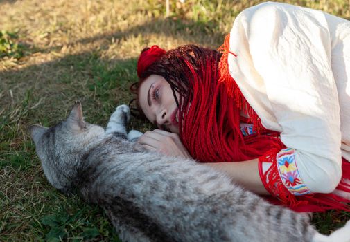 Young woman with scarlet dreadlocks in national dress lying on the grass and playing with the cat. Outdoors portrait