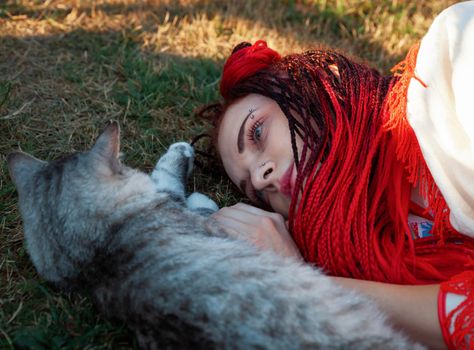 Young woman with scarlet dreadlocks in national dress lying on the grass and playing with the cat. Outdoors portrait