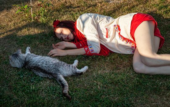 Young woman with scarlet dreadlocks in national dress lying on the grass and playing with the cat. Outdoors portrait